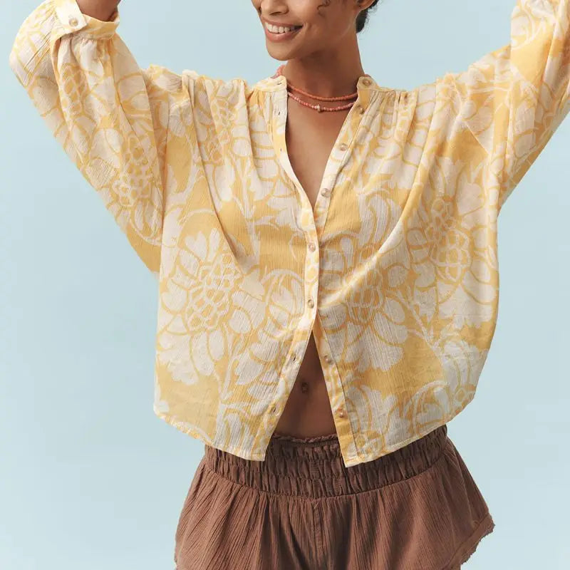 Woman wearing a yellow floral blouse and brown pants against a light blue background
