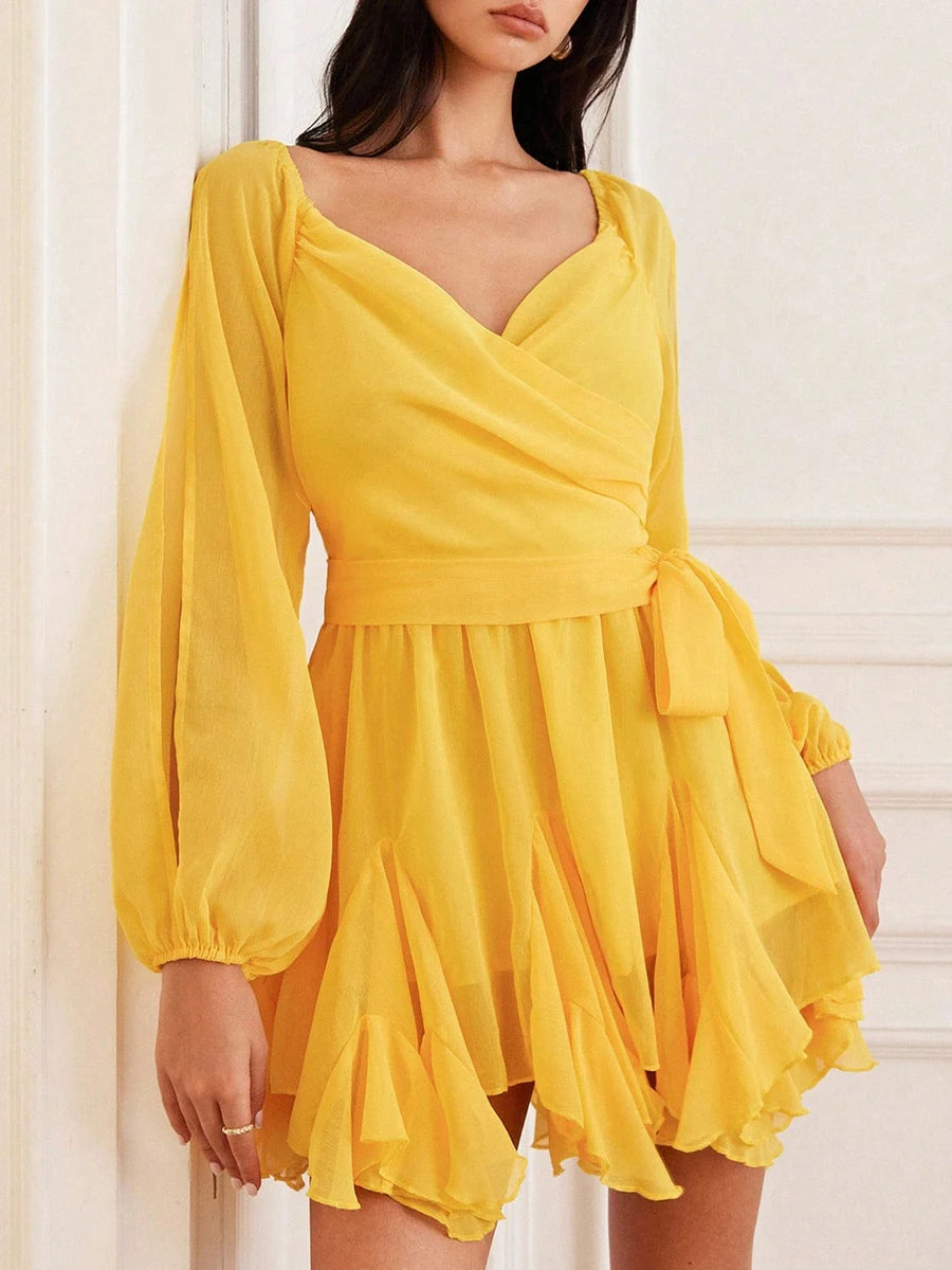 Yellow ruffle mini dress with long sleeves and a tie waist detail, worn by a person against a white background