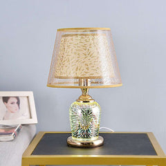  Golden Atelier 3D fireworks table lamp with a glass enclosure filled with intricate lighting elements that create a 3D fireworks effect. The lamp has a base with up-down lighting controls, displayed on a table in a living room setting. Rice White