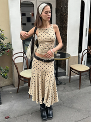 Woman in a polka dot dress standing on a sidewalk with chairs and a table in the background.