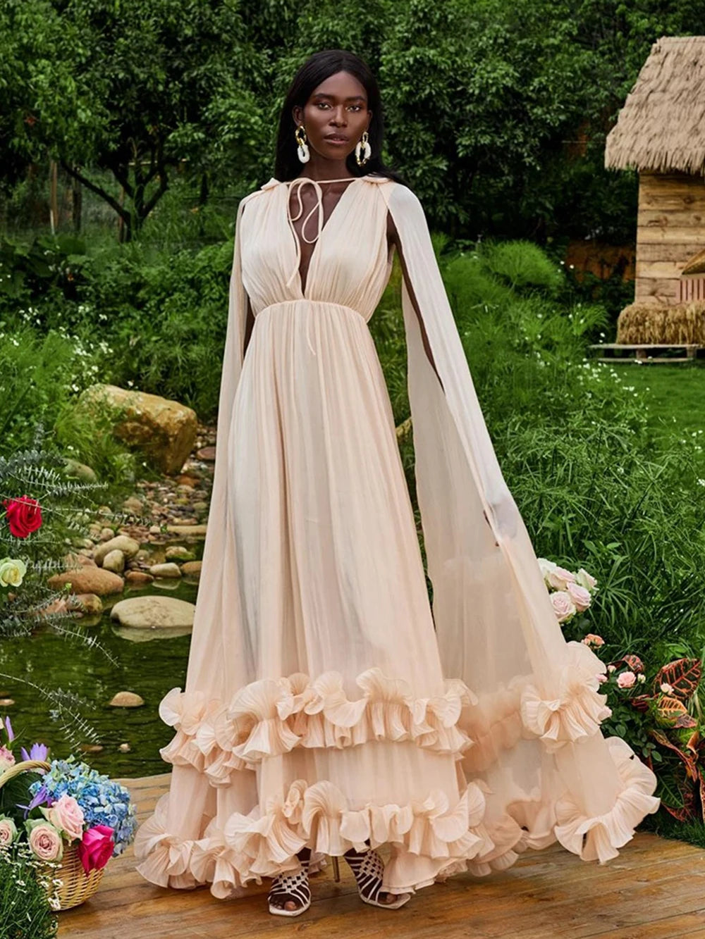 Woman in a long, flowing beige dress with ruffled details standing in a garden setting.