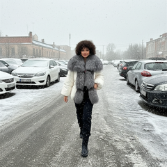 Person wearing a fur vest walking in a snowy street with cars and buildings in the background.
