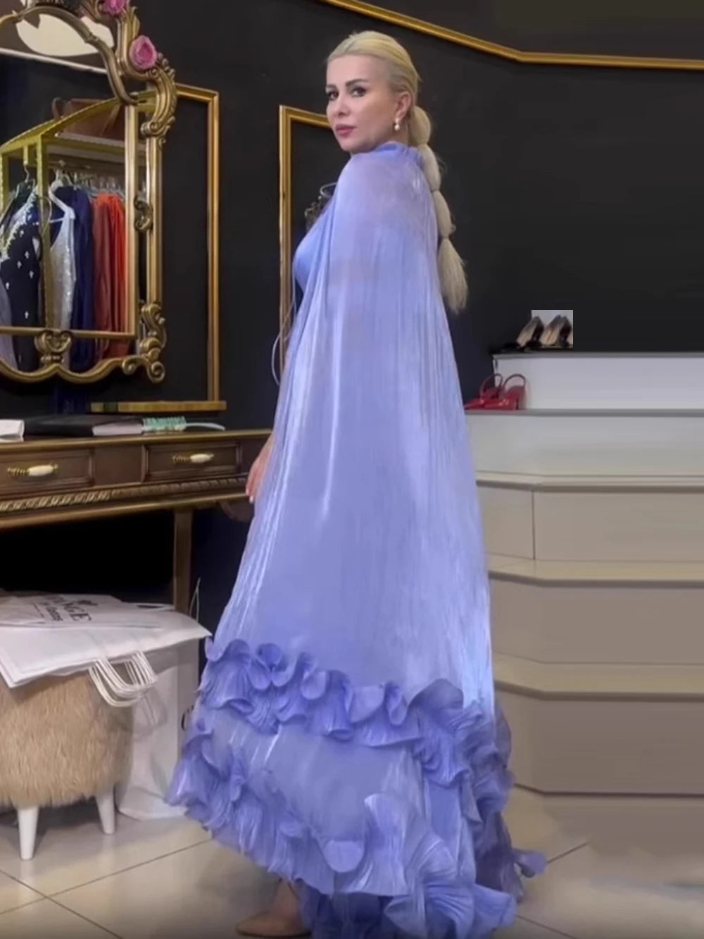 Woman wearing a long, light blue dress with ruffled layers in a room with mirrors and clothing racks.