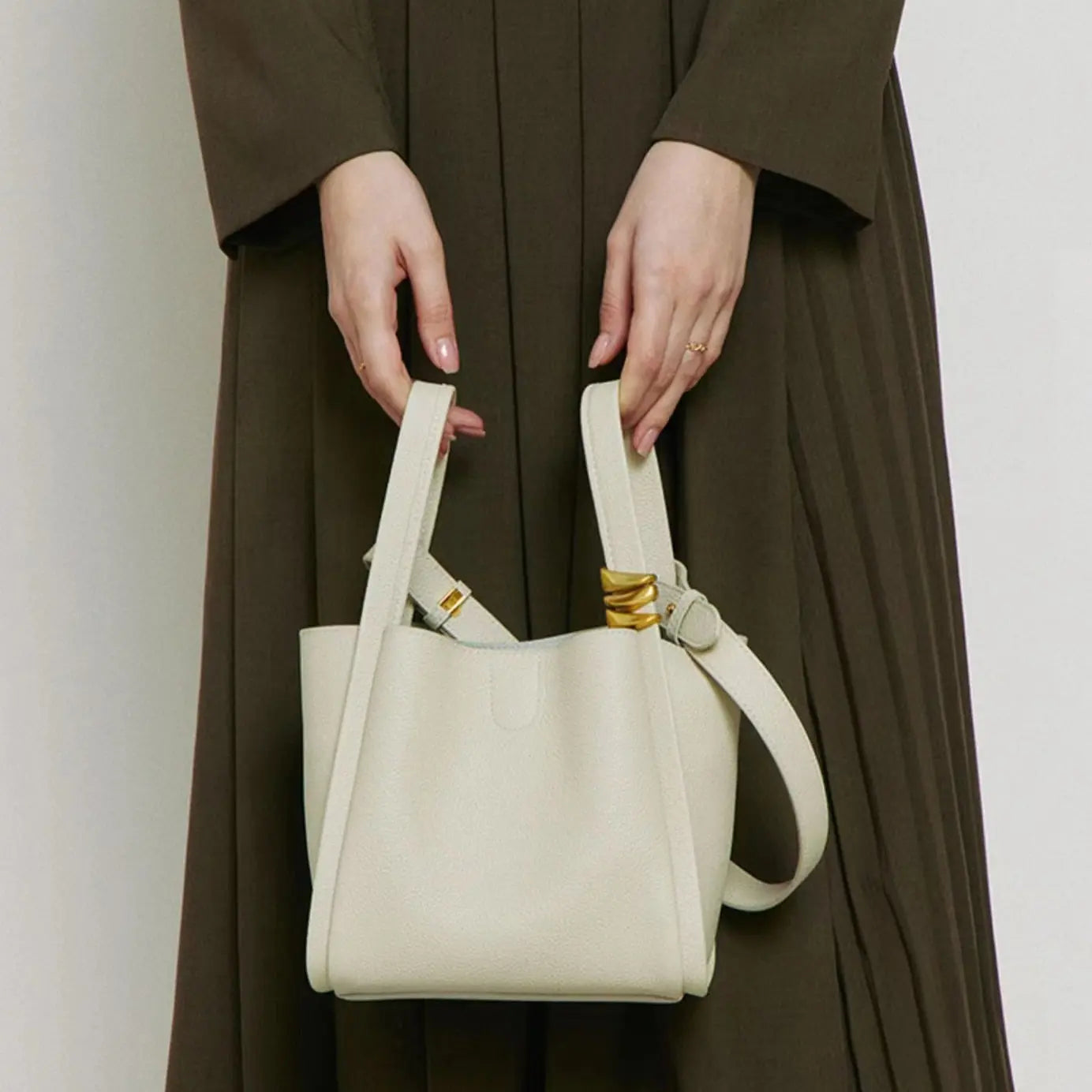 A faux leather bucket crossbody bag in beige colour with an adjustable strap and drawstring closure hold in the hand by a women model.