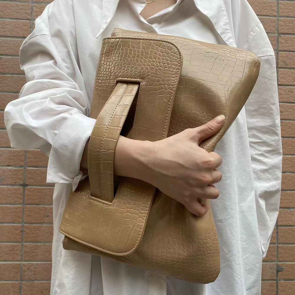 Person holding a beige crocodile-patterned clutch against a brick wall.