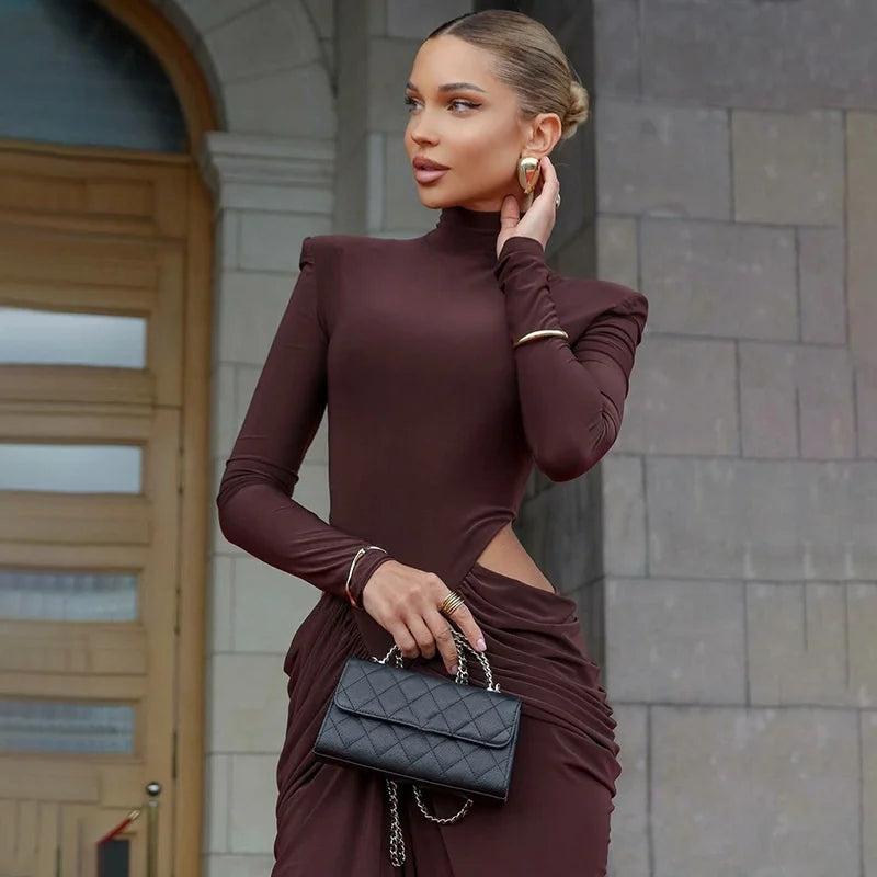 Woman in a burgundy dress holding a black handbag against a stone wall.