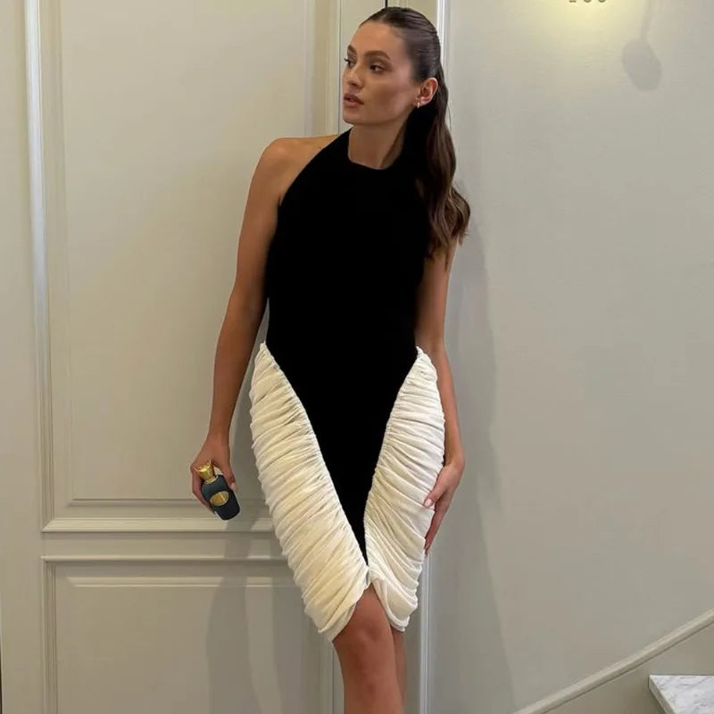 Woman wearing a black and white dress with a pleated skirt, holding a clutch, against a neutral wall.