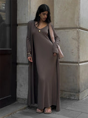 Woman wearing a long brown dress and cardigan standing against a stone wall.
