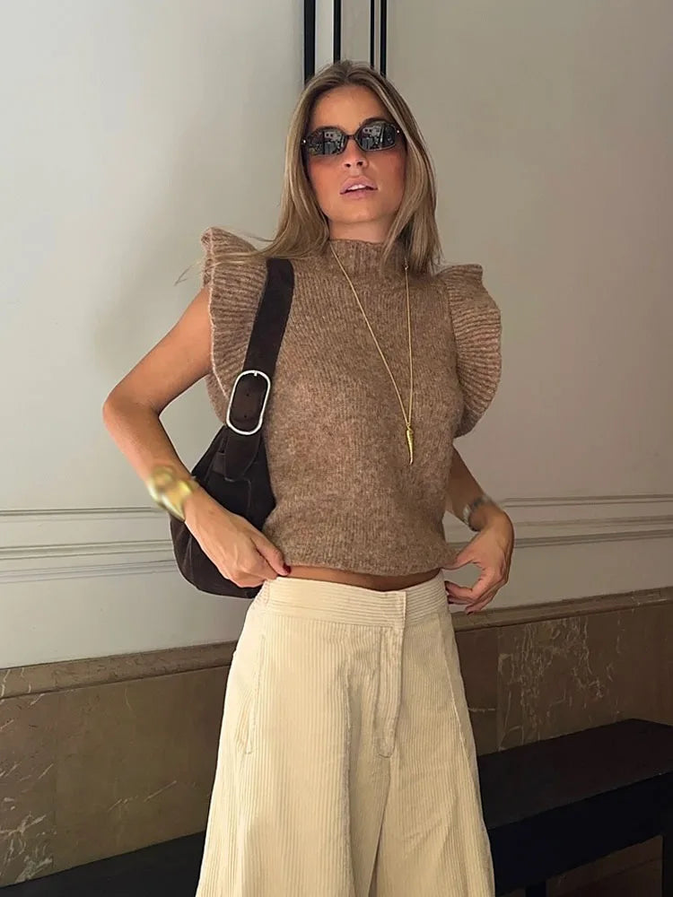 Woman wearing a brown sleeveless sweater and beige pants indoors.