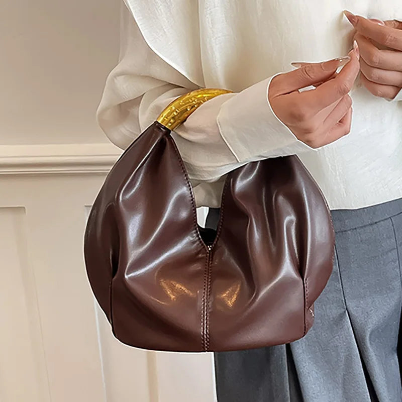 Brown soft PU leather dumpling shaped handbag with a textured gold metal handle, held by a hand.