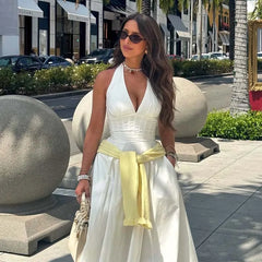 Woman in a white dress with a yellow belt standing on a sidewalk with palm trees and shops in the background.