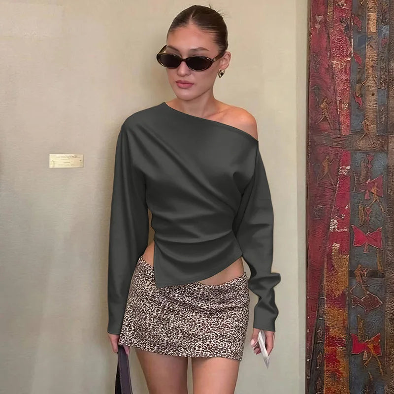 Woman wearing a dark gray off-shoulder top and patterned skirt standing against a plain wall.