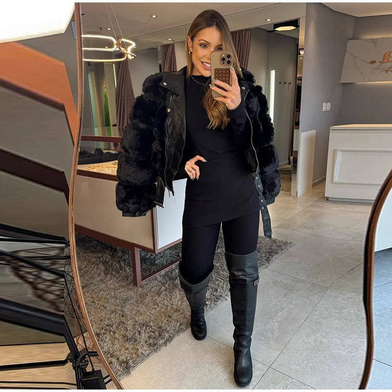 Woman taking a mirror selfie wearing a black outfit with a fur-lined coat in a modern interior setting.