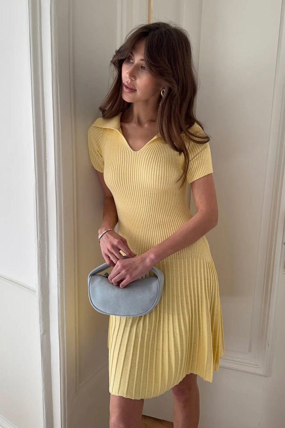 Woman in a yellow dress holding a blue clutch against a neutral wall.