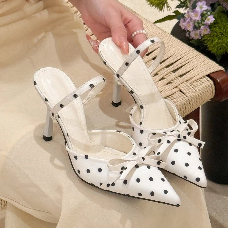 White high-heeled shoes with black polka dots and bows held by a hand.