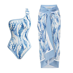 One-shoulder one-piece swimsuit with blue scale print and a metal ring detail, paired with a wrap-style skirt.