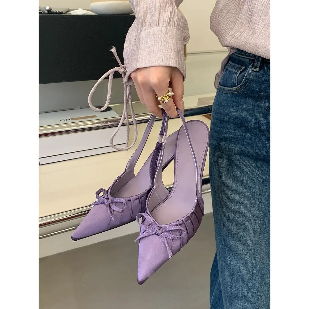Purple heeled shoes held by a person wearing a beige sweater and blue jeans.