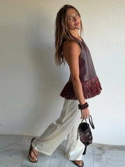 Woman wearing a sleeveless brown top with fringe and white pants, holding a black handbag.
