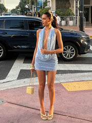 Woman in a light blue dress standing on a city street with cars and pedestrians in the background.