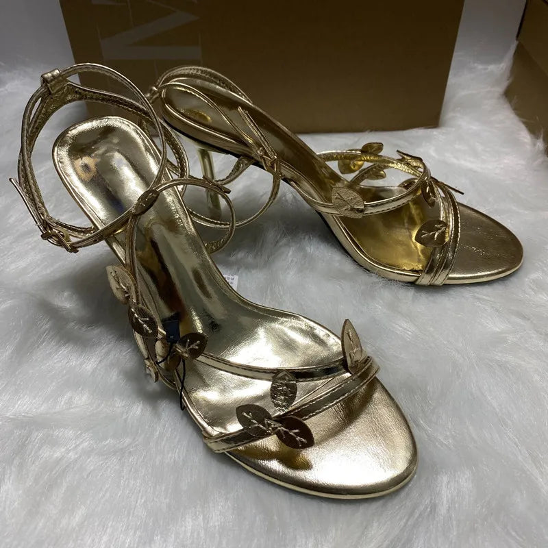 Gold high-heeled sandals on a white surface with a brown box in the background