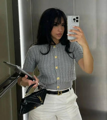 Woman taking a mirror selfie wearing a gray cardigan with gold buttons, white pants, and holding a black handbag.