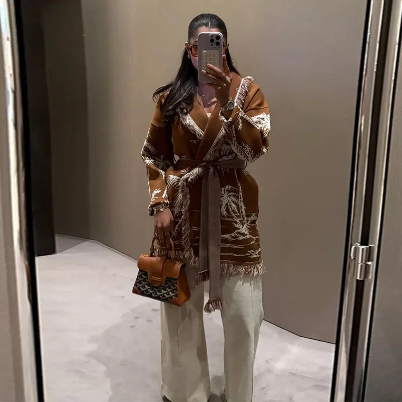 Person wearing a brown patterned coat and white pants, holding a phone and a bag, standing in front of a mirror.