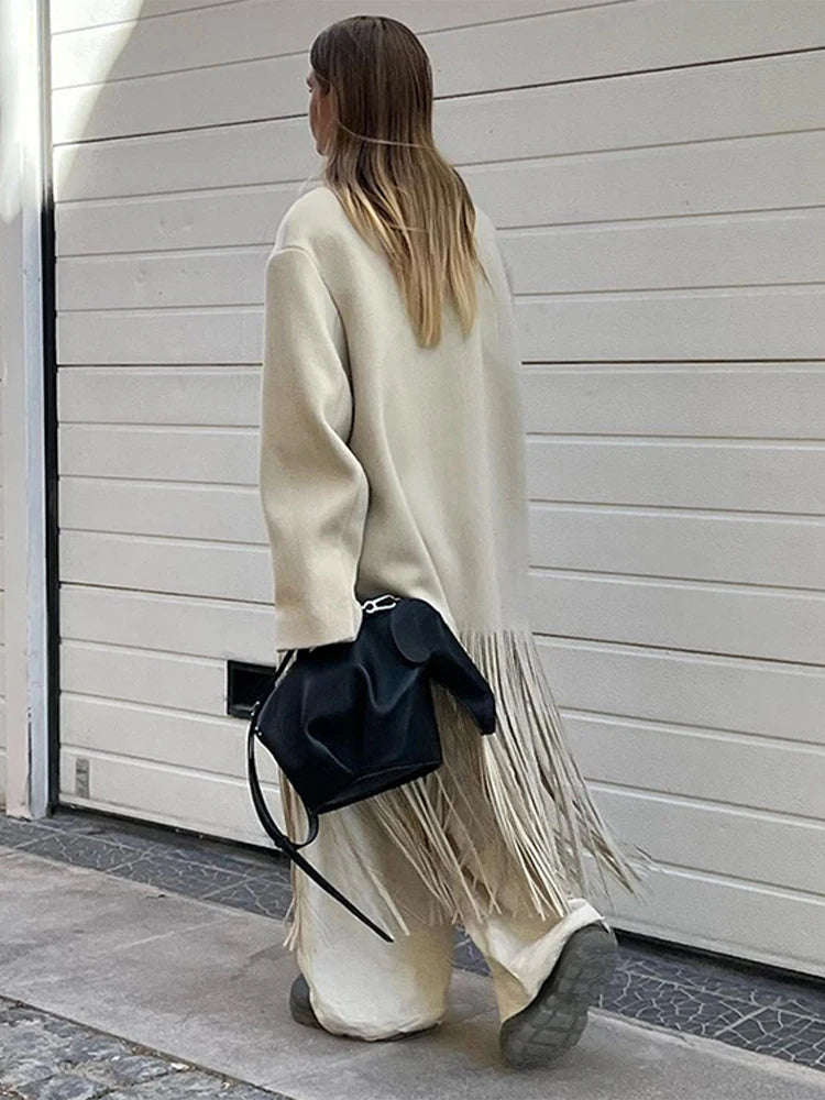 Person wearing a long cream coat with a black bag, walking past a white wall.