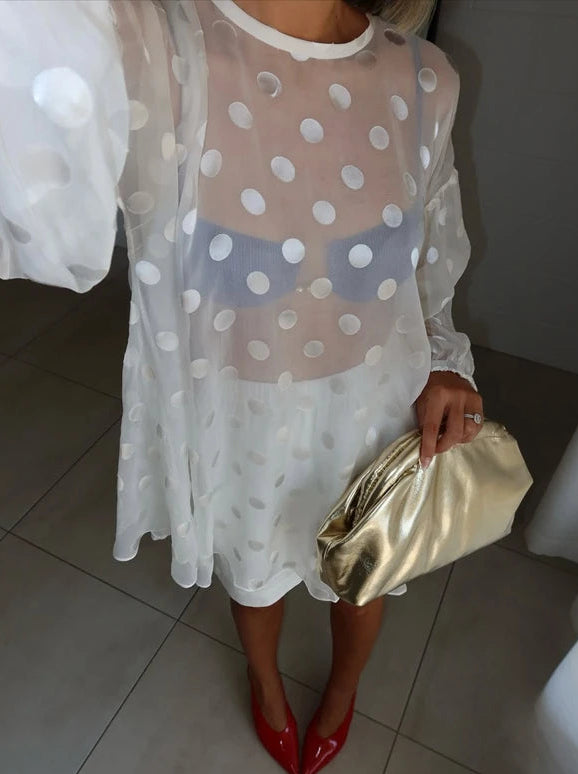 Person wearing a white polka dot dress with sheer overlay, holding a gold clutch, and wearing red shoes.