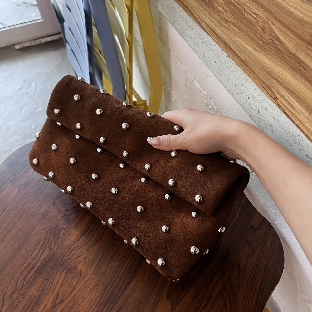 Brown clutch bag with silver studs held by a hand on a wooden surface.