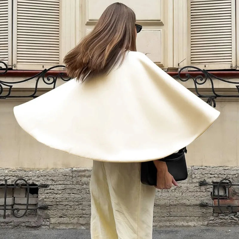 Person wearing a beige cape and holding a black bag on a street with stone walls and shutters.