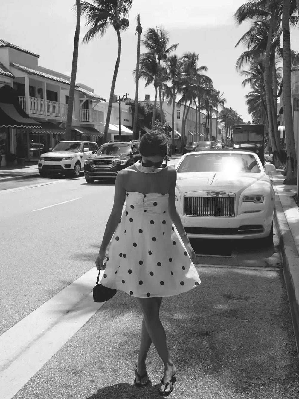Woman in a polka dot dress walking on a street with palm trees and a luxury car in the background.