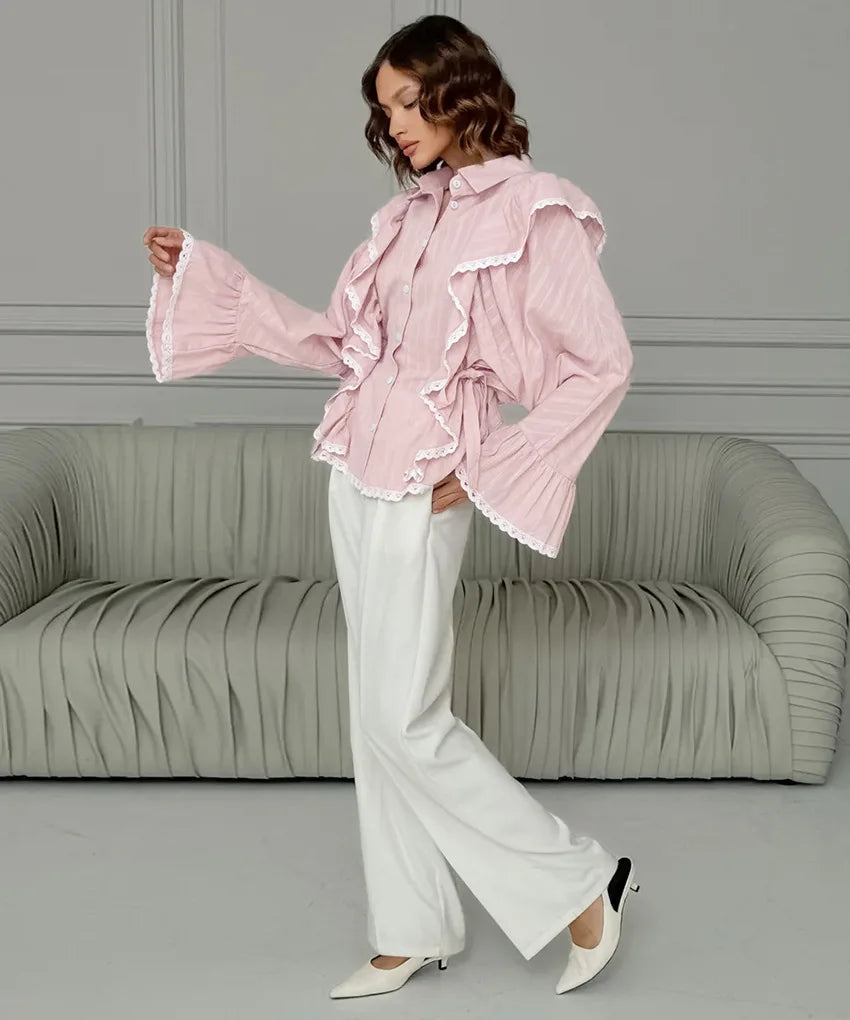 Woman wearing a pink ruffled blouse and white pants in a room with a gray sofa.