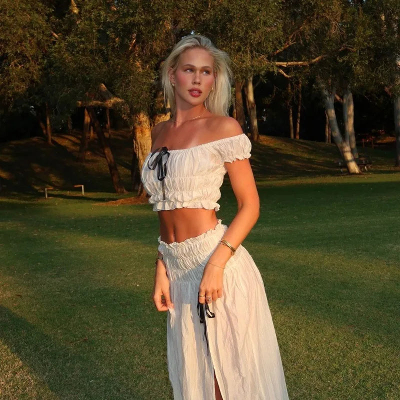 Woman in a white off-shoulder top and skirt standing in a grassy outdoor area.