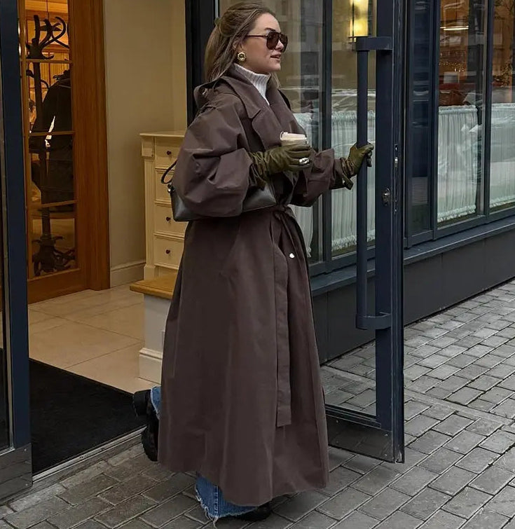 Woman in a long brown coat standing outside a store holding a coffee cup.