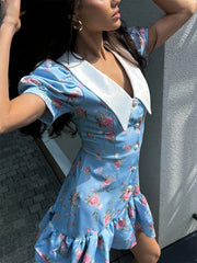 Woman wearing a blue floral dress with a white collar against a gray wall.