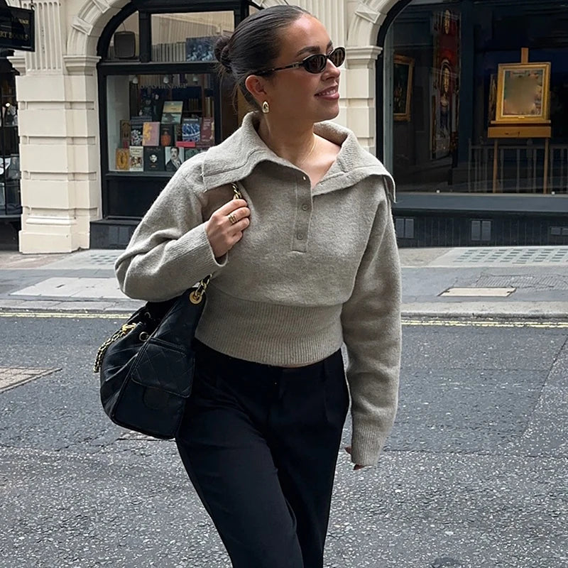 A woman wearing a knitted polo sweater, leaning casually near a storefront with tailored trousers and sunglasses