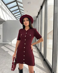 Woman in a burgundy dress and hat standing in a modern indoor setting with large windows.