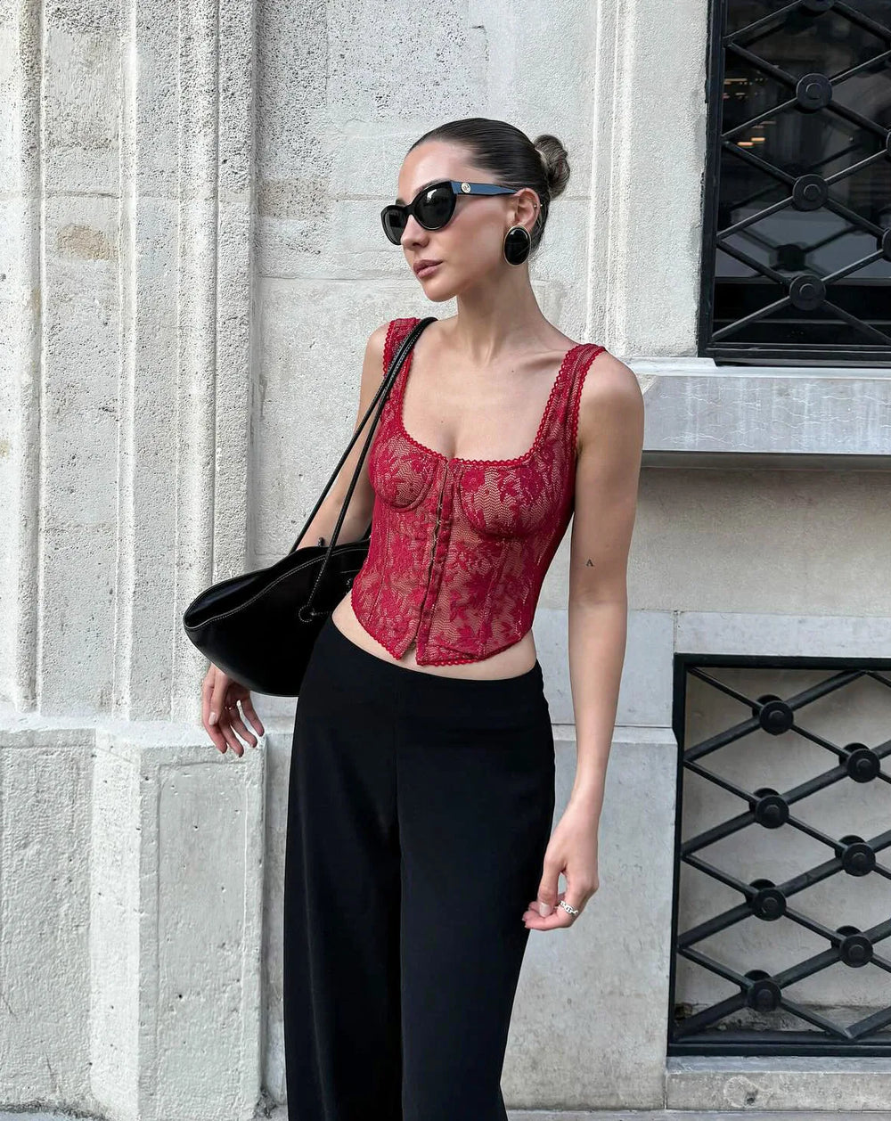 Woman wearing a red lace top and black pants standing against a stone wall.