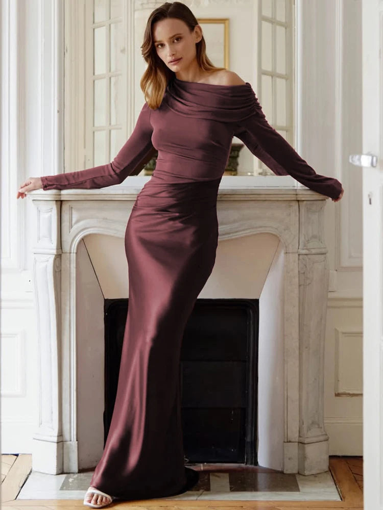 Woman in a long, dark purple off-shoulder dress standing in a room with a fireplace.