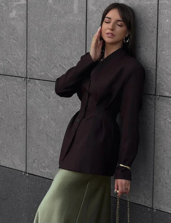 Woman wearing a black blazer and green pants against a gray tiled wall.