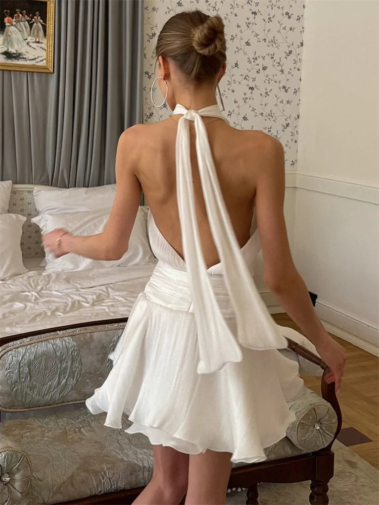 Woman in a white dress with a halter neck and open back, standing in a room with decorative elements.