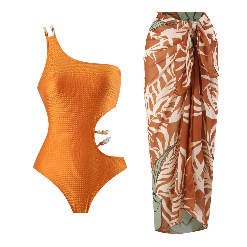 Orange textured one-shoulder swimsuit with cutout details featuring beaded embellishments, paired with a brown and white tropical leaf print sarong.