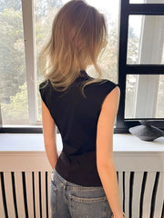 Person wearing a black sleeveless top and jeans standing by a window.
