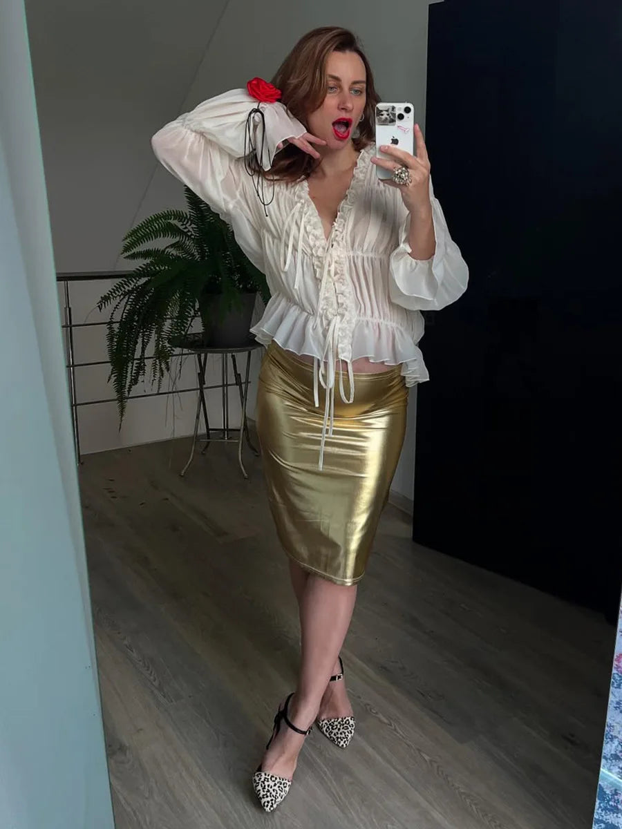 Woman taking a mirror selfie wearing a white blouse and gold skirt indoors.