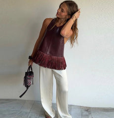Woman wearing a brown fringed top and white pants, holding a black handbag with a pattern.
