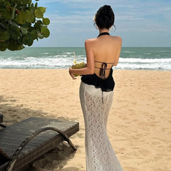 Woman in a black top and silver skirt standing on a beach holding a coconut drink.