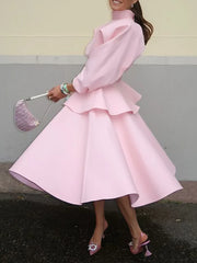 Woman wearing a pink dress with ruffled details, holding a matching handbag against a neutral background.