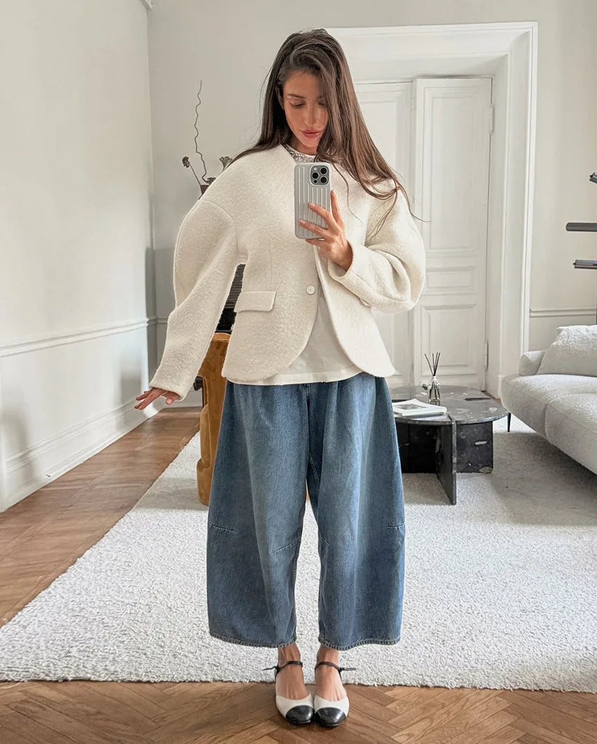 Woman taking a mirror selfie in a stylish outfit with a white blazer and blue pants in a modern living room.