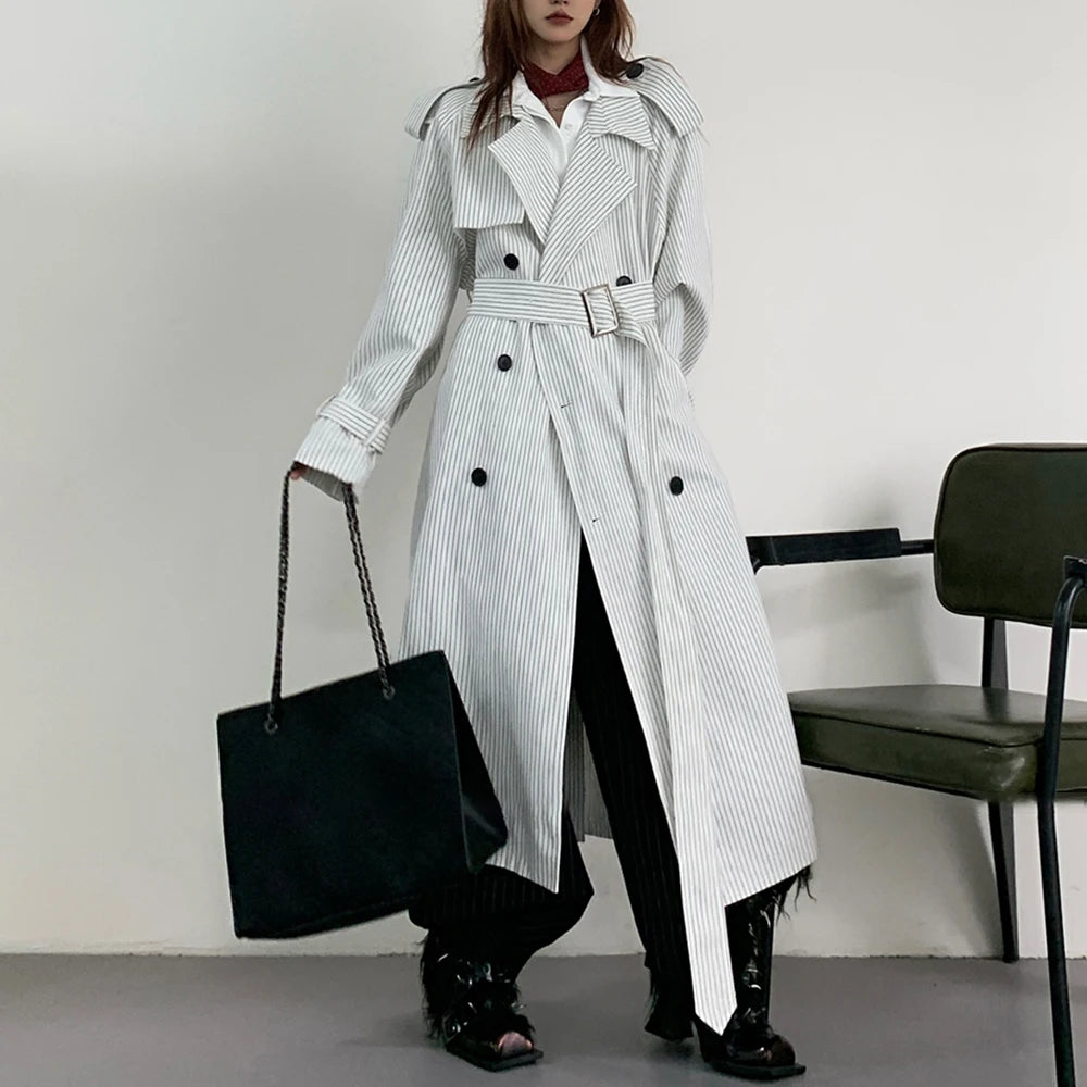 Person wearing a long black and white striped trench coat holding a black handbag against a plain background.