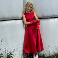 Woman in a red dress and gloves leaning against a white wall with grass at the base.